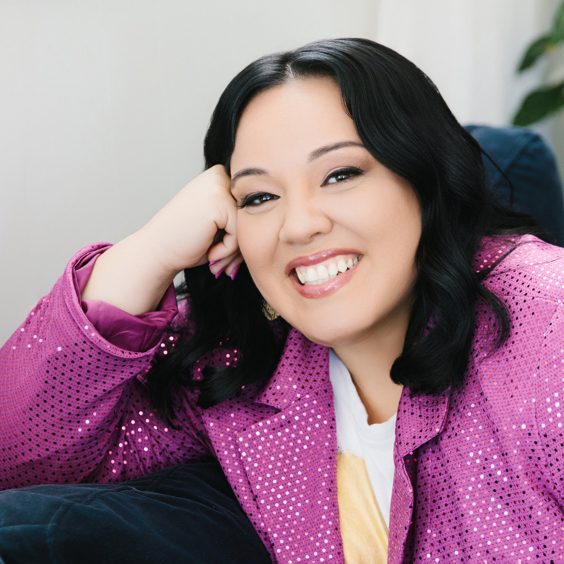 Smiling woman with black hair wearing a sparkly purple jacket resting her head on her hand.