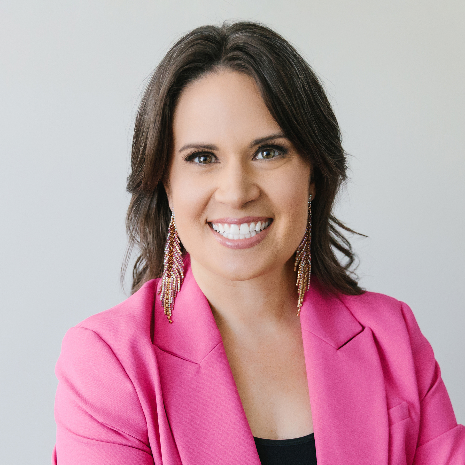 Smiling woman with shoulder-length brown hair wearing a bright pink blazer and long beaded earrings.