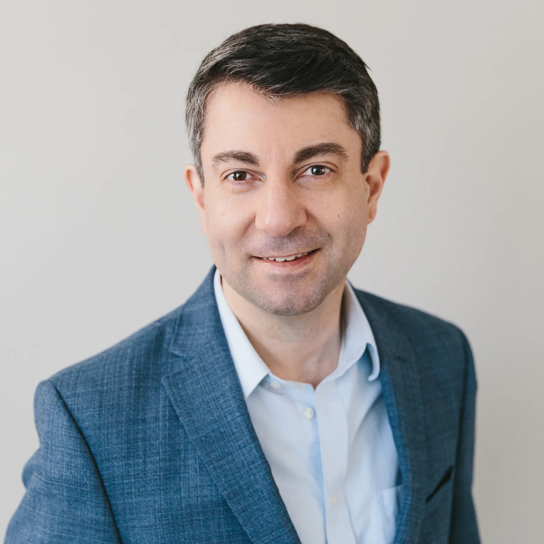 Smiling man with short dark hair wearing a blue suit jacket and light blue shirt against a plain background.