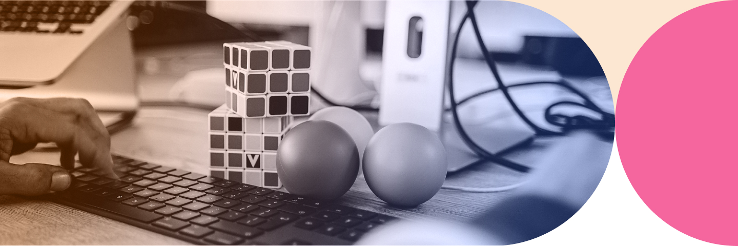 Hand typing on a black keyboard next to stacked Rubik's cubes and three balls on a wooden desk with computer equipment in the background.