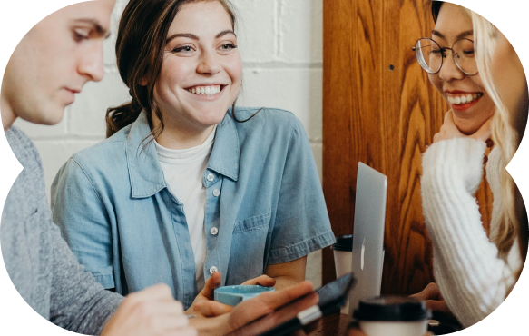 Three young adults smiling and engaging in a casual conversation around a table with laptops and coffee cups.
