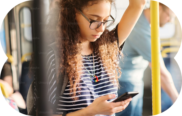 Young woman with curly hair and glasses looking at her phone while standing on public transport and holding a yellow pole.