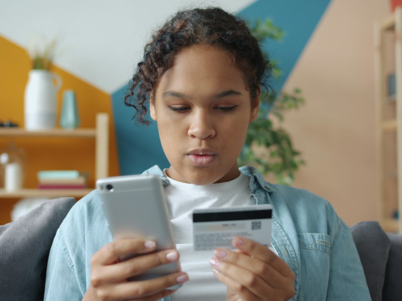 A young woman using a phone to purchase online. She is holding a debit card on one hand and a phone in the ther. She has a concentrated expression.