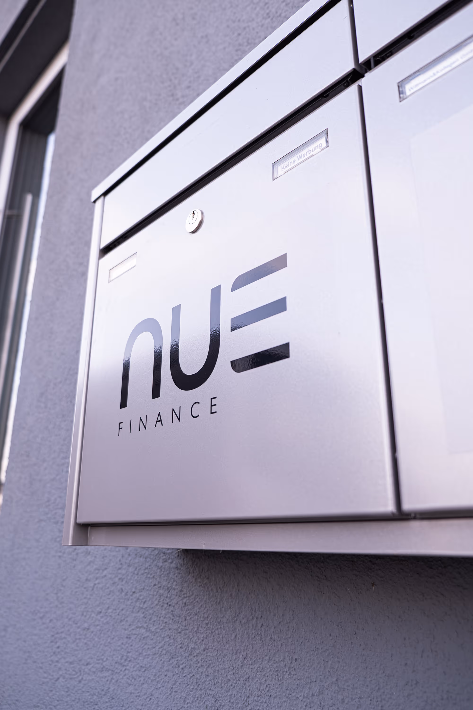 Metallbriefkasten mit dem Schriftzug 'NUE FINANCE' an einer grauen Wand montiert.
