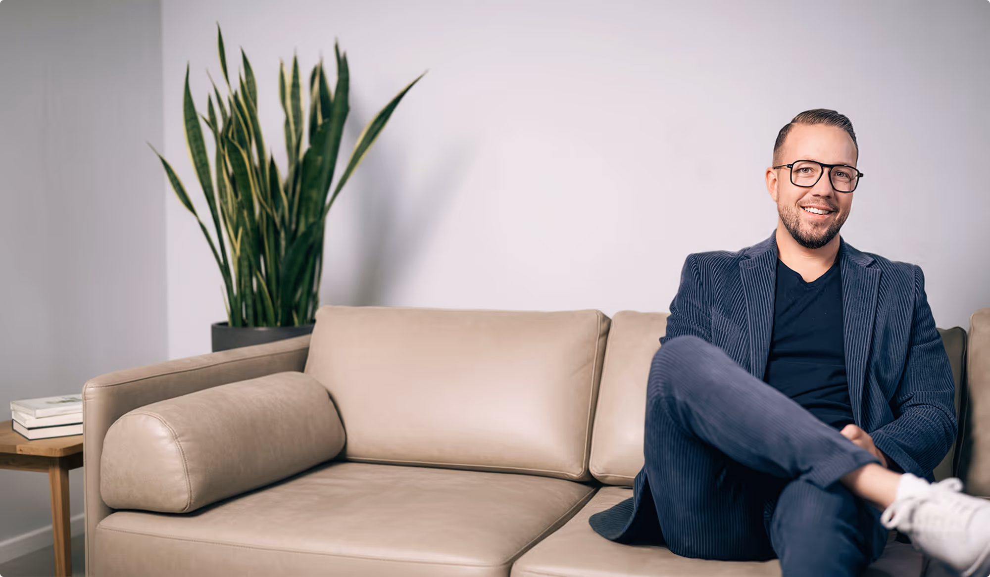 NUE-Geschäftsführer Pascal Baumüller sitzt lächelnd mit verschränkten Beinen auf einem hellen Ledersofa, im Hintergrund eine hohe grüne Pflanze.