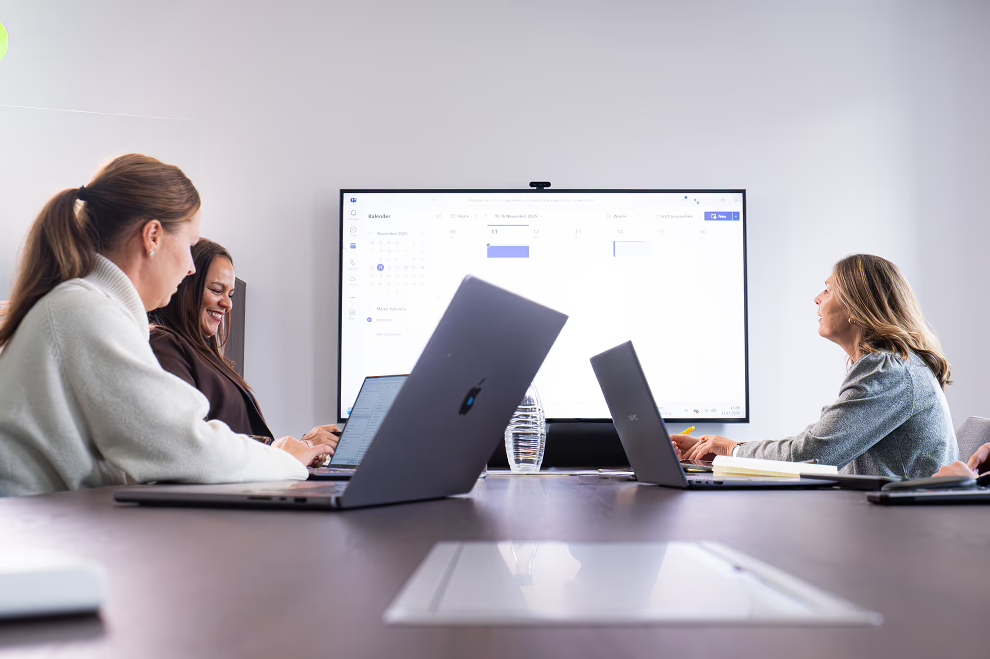 Drei Frauen sitzen mit Laptops an einem Konferenztisch vor einem großen Bildschirm mit einem Online-Kalender.