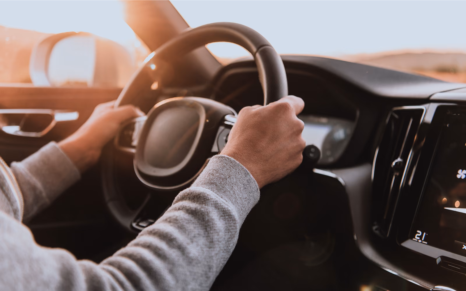 Hände halten das Lenkrad eines Autos, durch die Frontscheibe fällt warmes Licht.