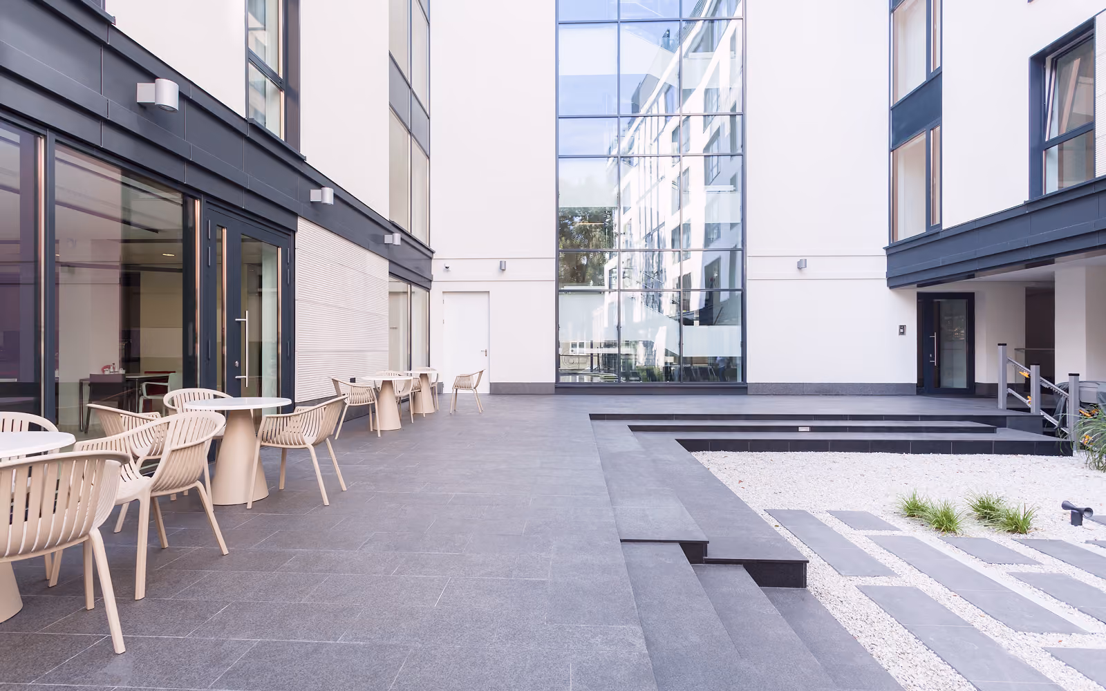 Moderner Gebäudekomplex mit Innenhof, Terrasse und Stühlen vor einer Glasfassade.