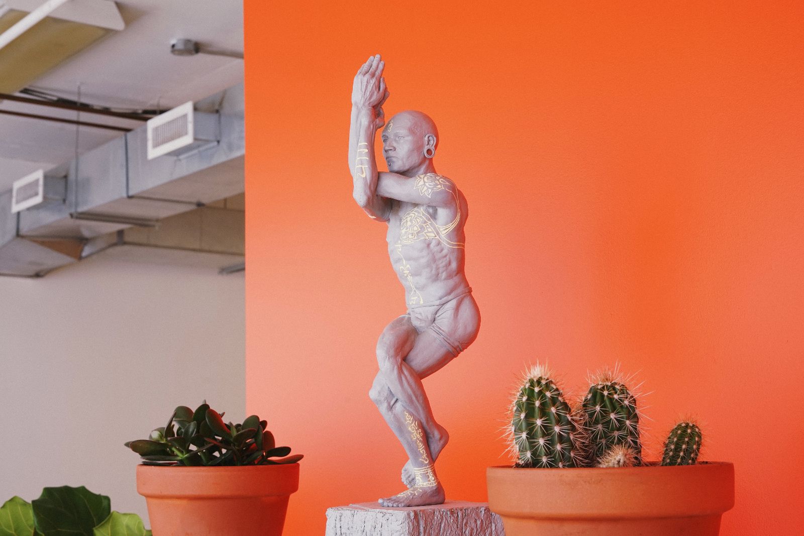 Clay sculpture of a tattooed man in a yoga pose between two terracotta pots with plants, against an orange wall.