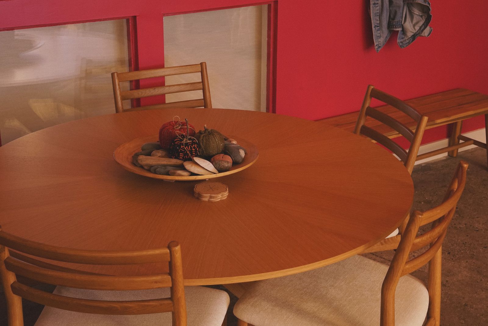 Round wooden dining table with four chairs and a decorative bowl of artificial pumpkins and stones, against a red wall with hanging jacket and wooden bench.