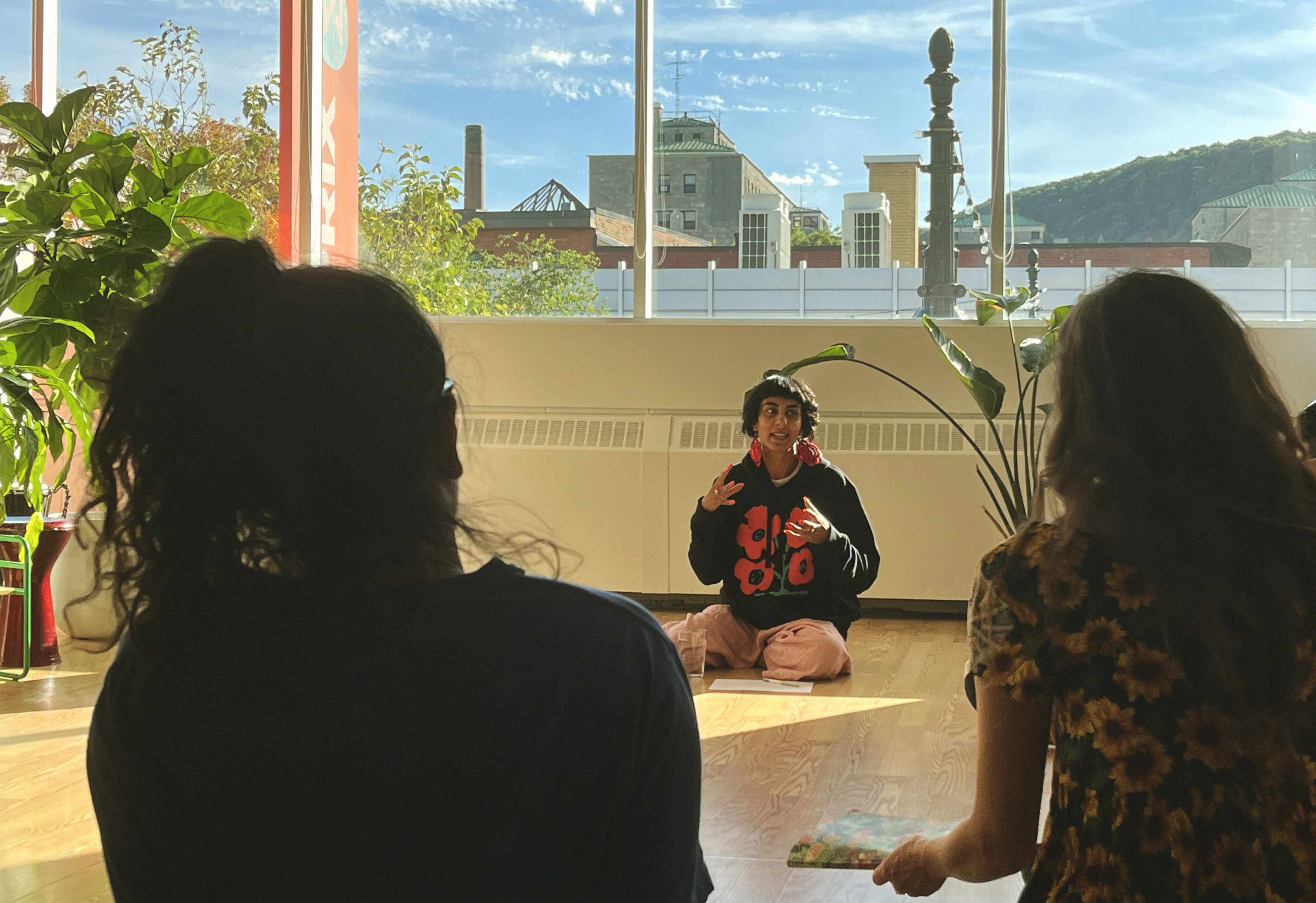 Person sitting cross-legged on the floor speaking to two seated listeners in a brightly lit room with large windows showing buildings and a hill outside.