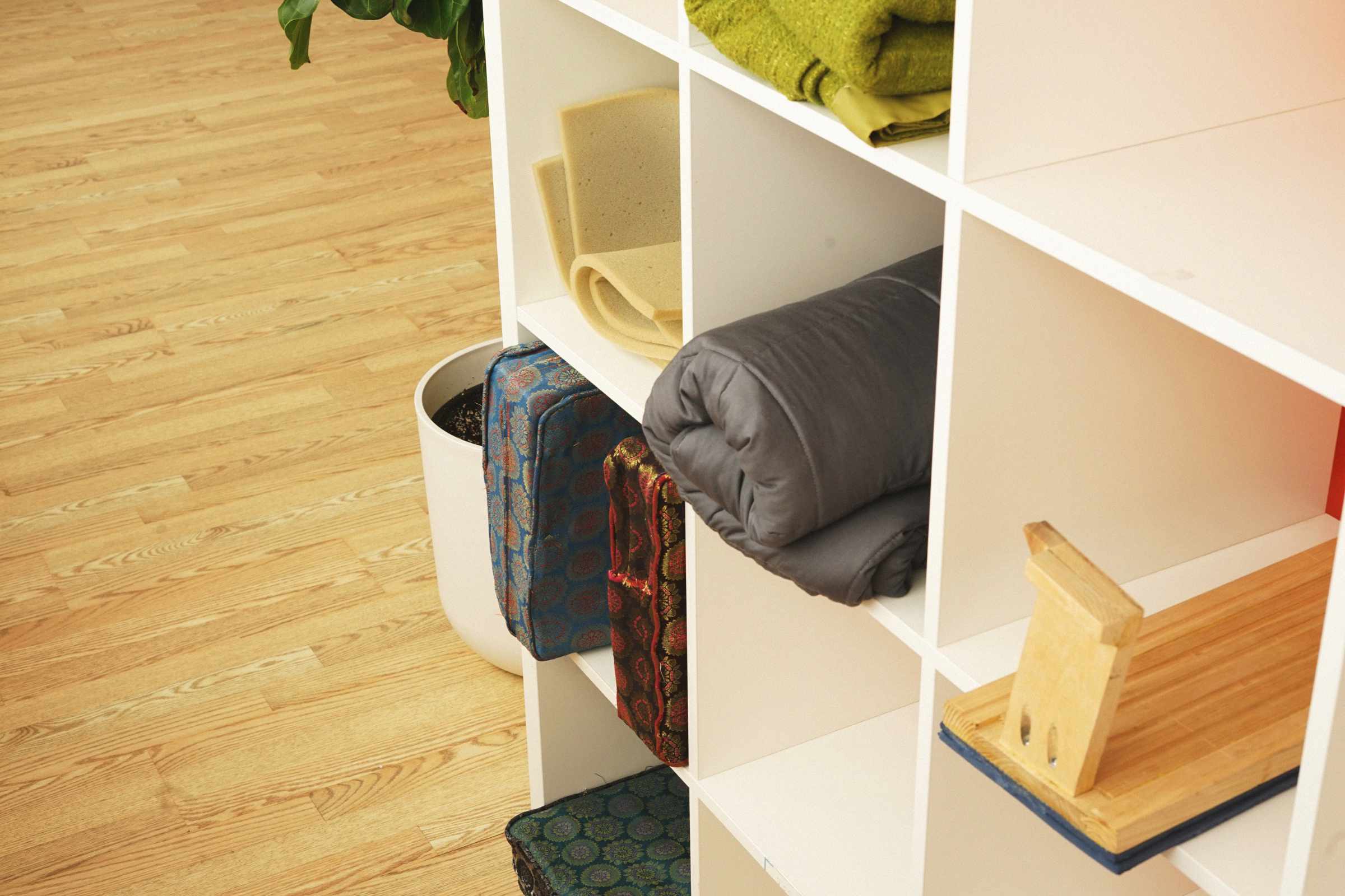 White cubby shelf with folded grey blanket, green folded textiles, rolled foam, patterned cushions, and a wooden book holder, next to a white plant pot on wooden floor.