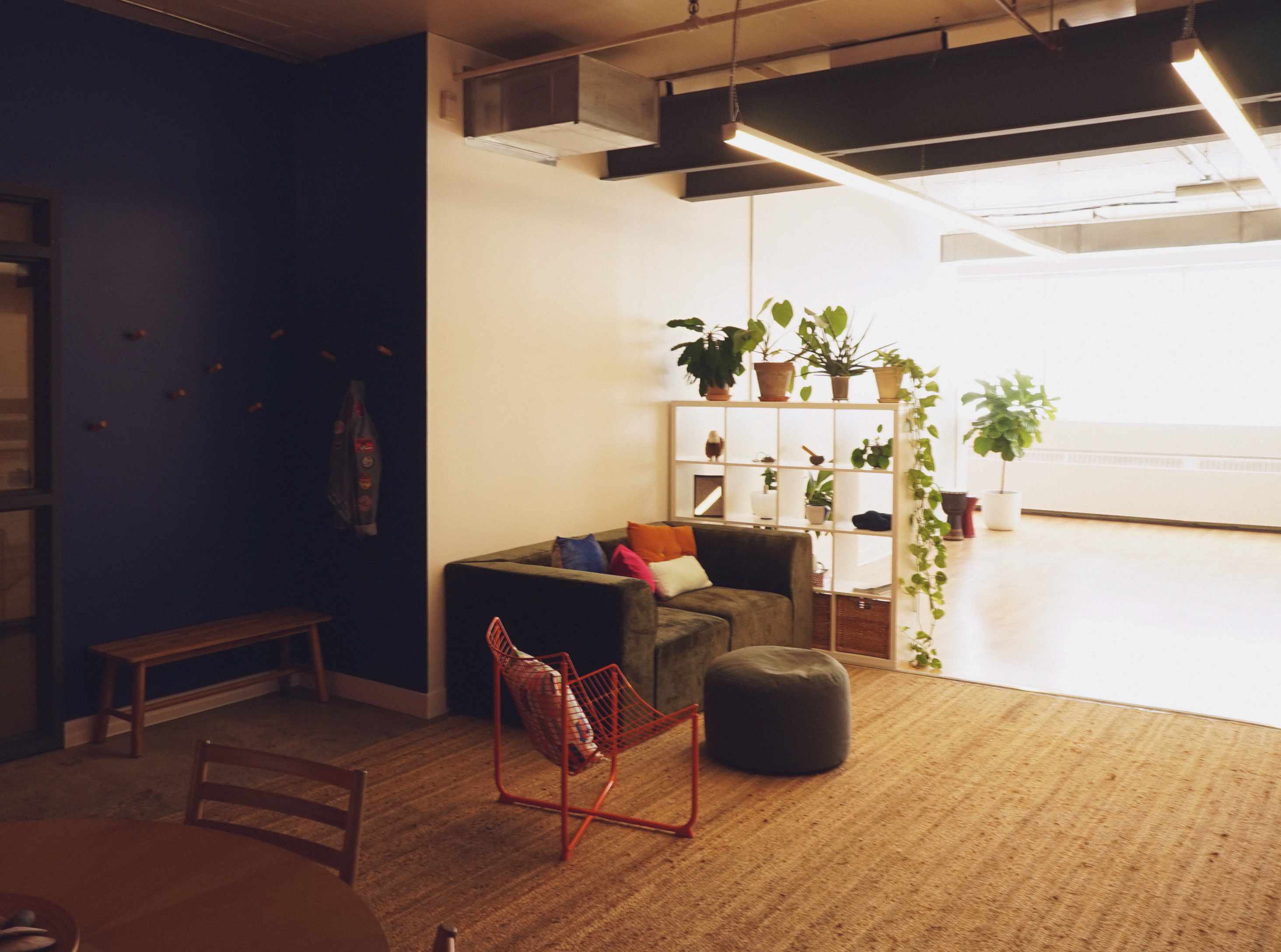 Cozy indoor seating area with a green couch, orange wire chair, pouf, wooden bench, and white shelving unit with potted plants.
