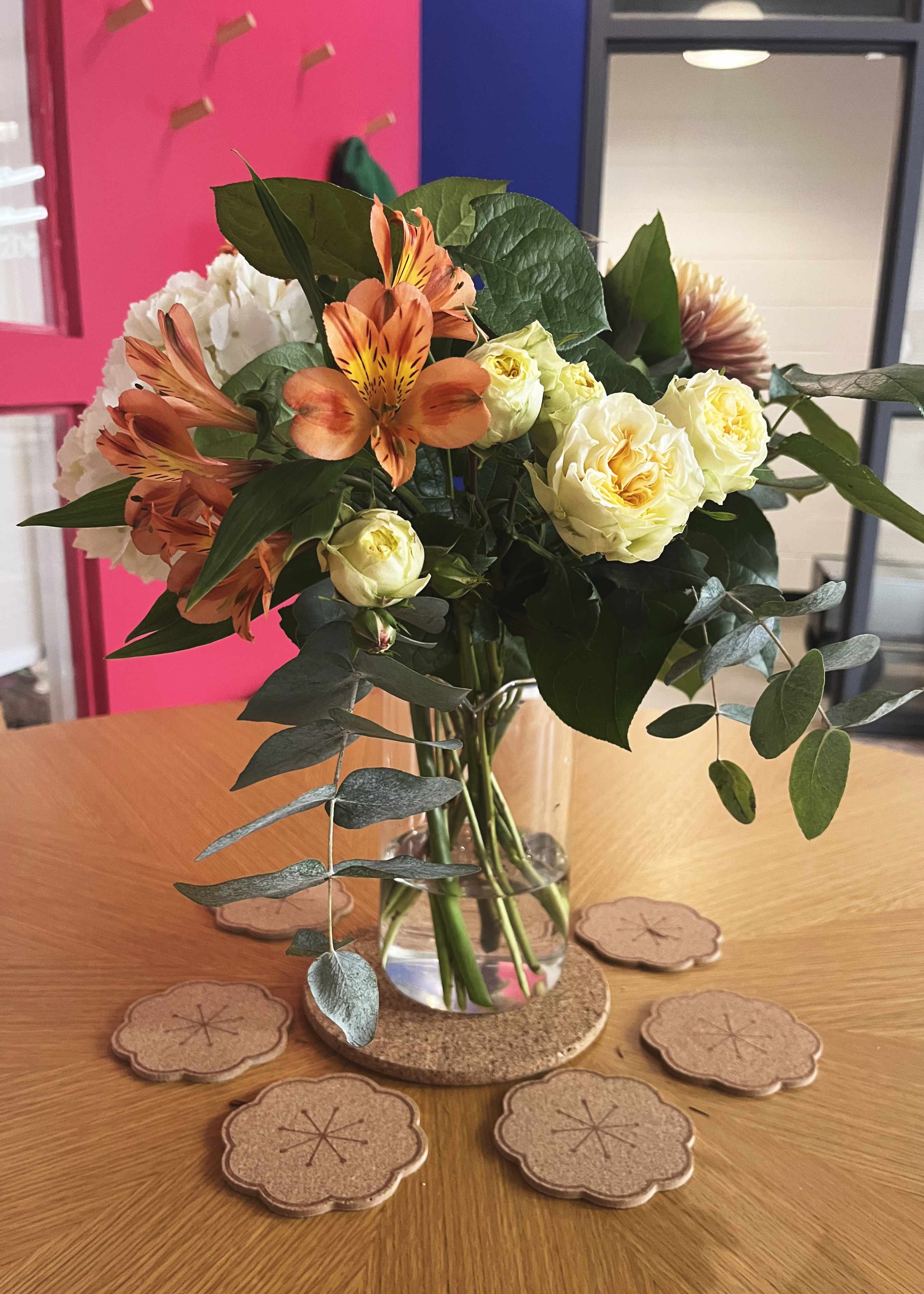 Vase with mixed flowers including orange Alstroemeria and cream roses on a wooden table with flower-shaped cork coasters.