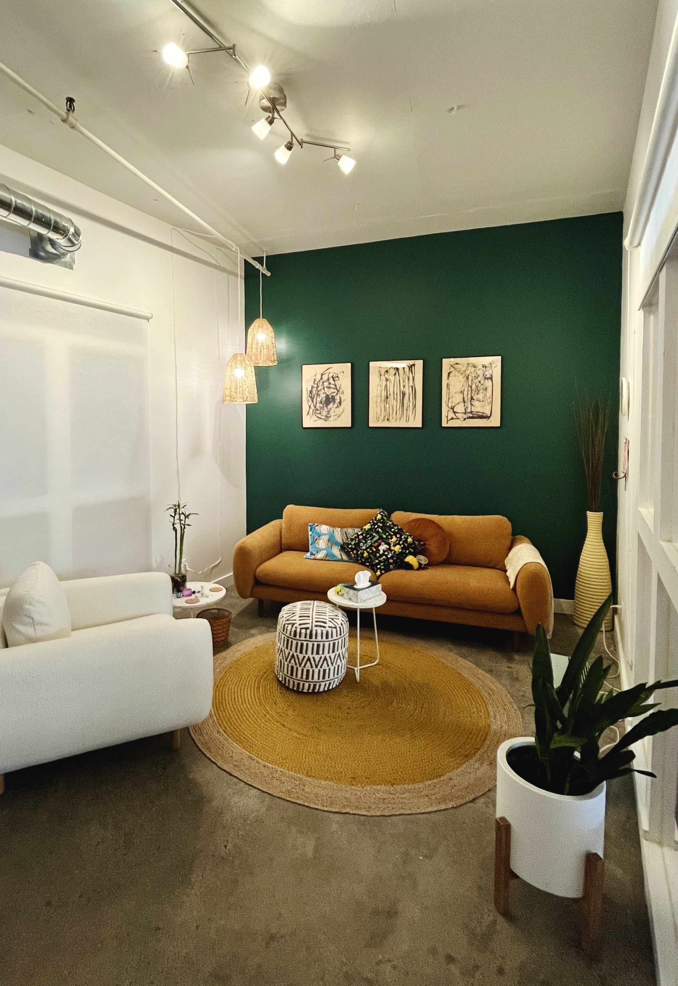 Cozy living room with a mustard yellow sofa against a dark green wall, white armchair, round mustard rug, patterned ottoman, and hanging woven pendant lights.