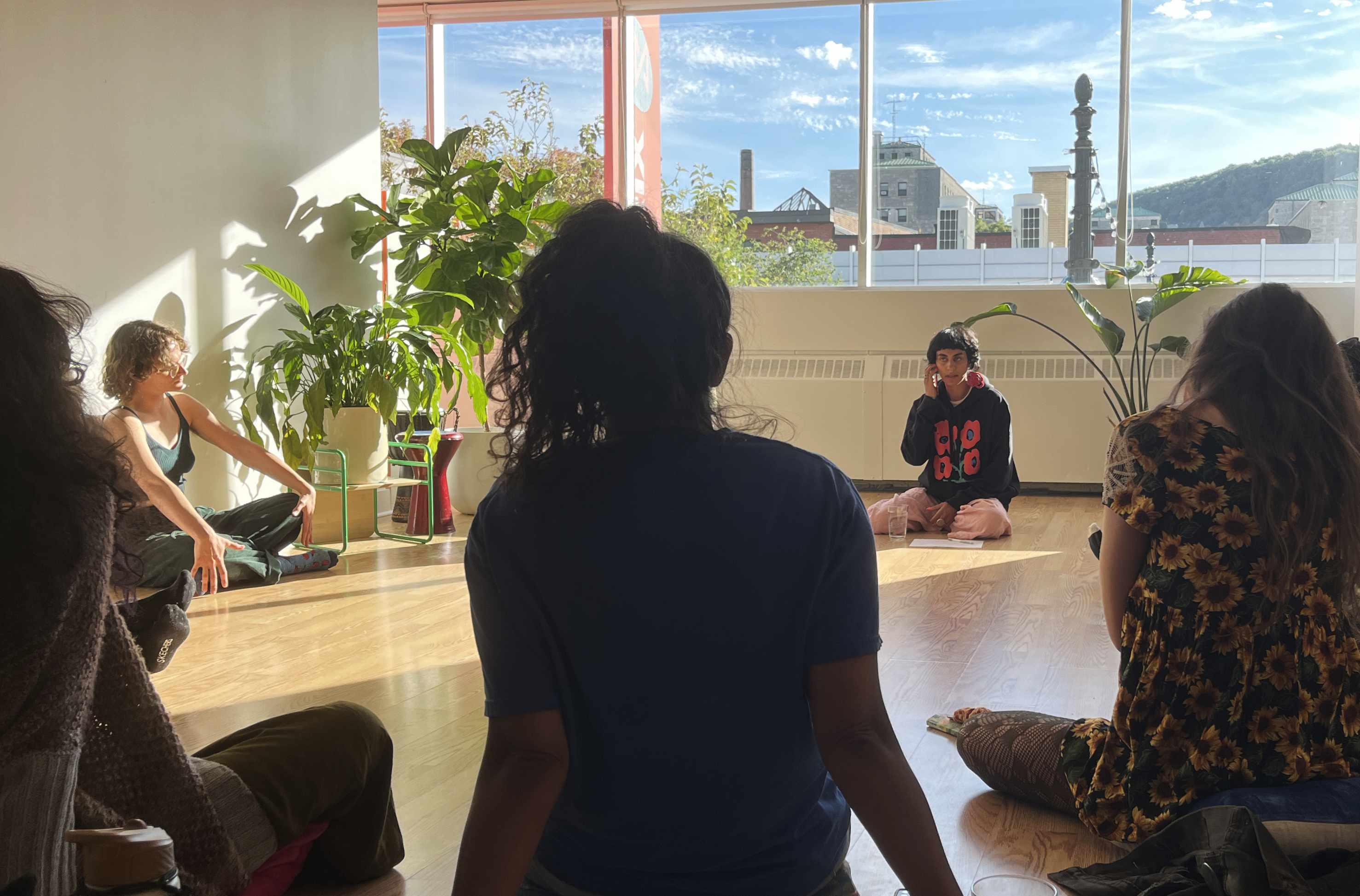 Group of people sitting on the floor in a sunlit room with large windows and indoor plants.