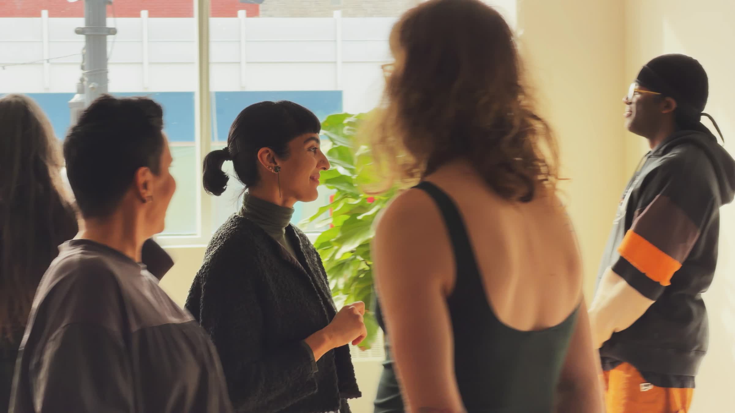Group of five people standing and talking indoors near a window and a green plant.