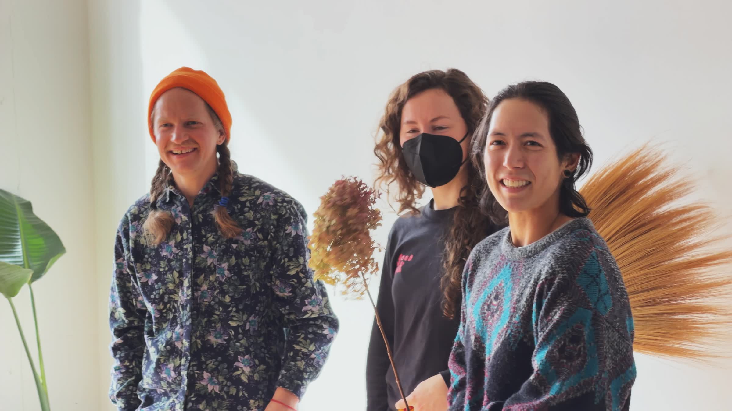 Three people smiling indoors, one wearing an orange beanie and floral shirt, another with curly hair and a black face mask, and the third wearing a patterned sweater holding dried flowers.