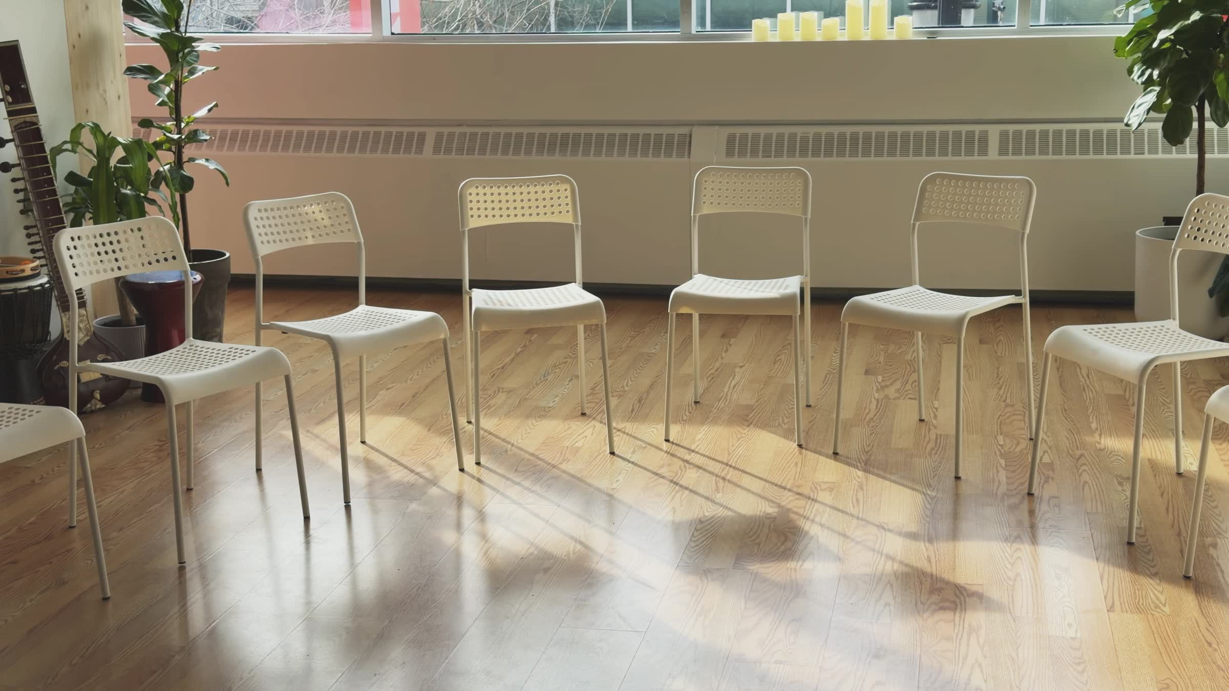Empty white perforated chairs arranged in a circle on a wooden floor with plants and musical instruments near a large window.