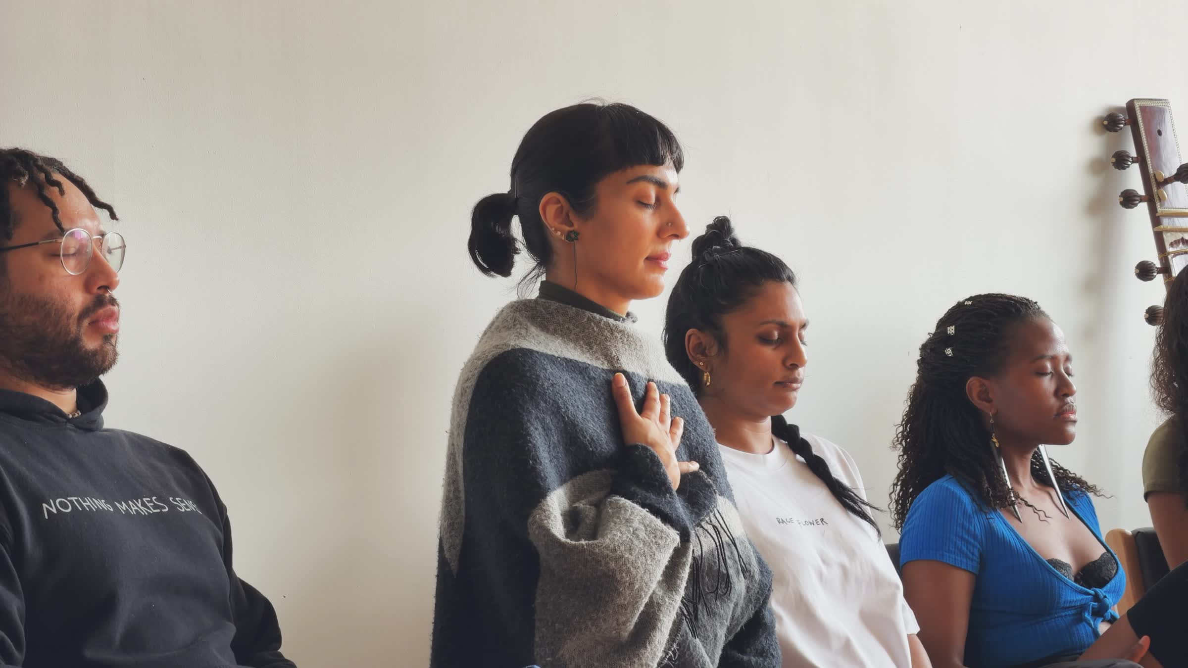 Four diverse people sitting in a row with eyes closed, practicing meditation or mindfulness indoors.