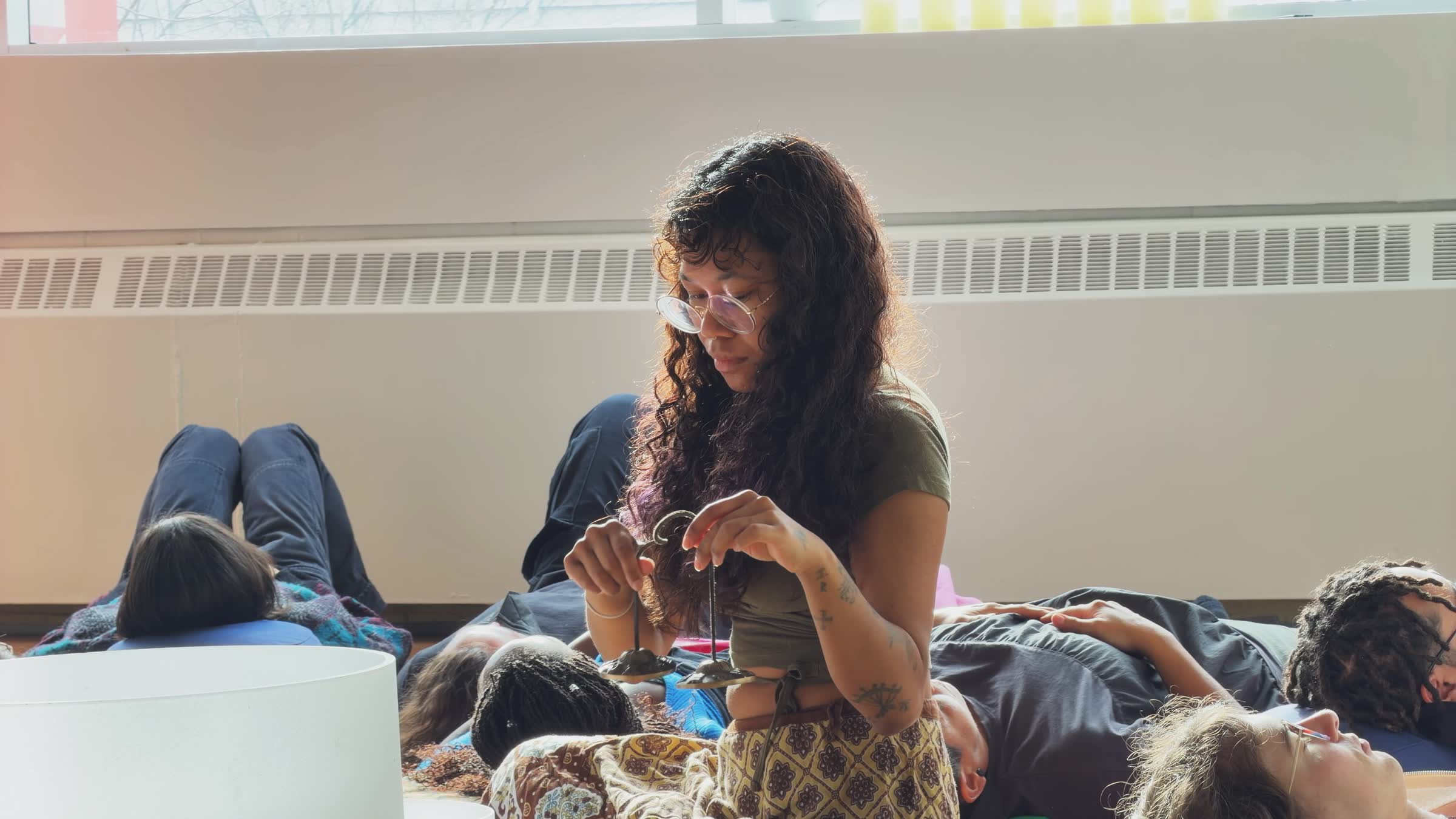 Young woman with curly hair and glasses holding small cymbals, with several people lying down resting behind her in a sunlit room.