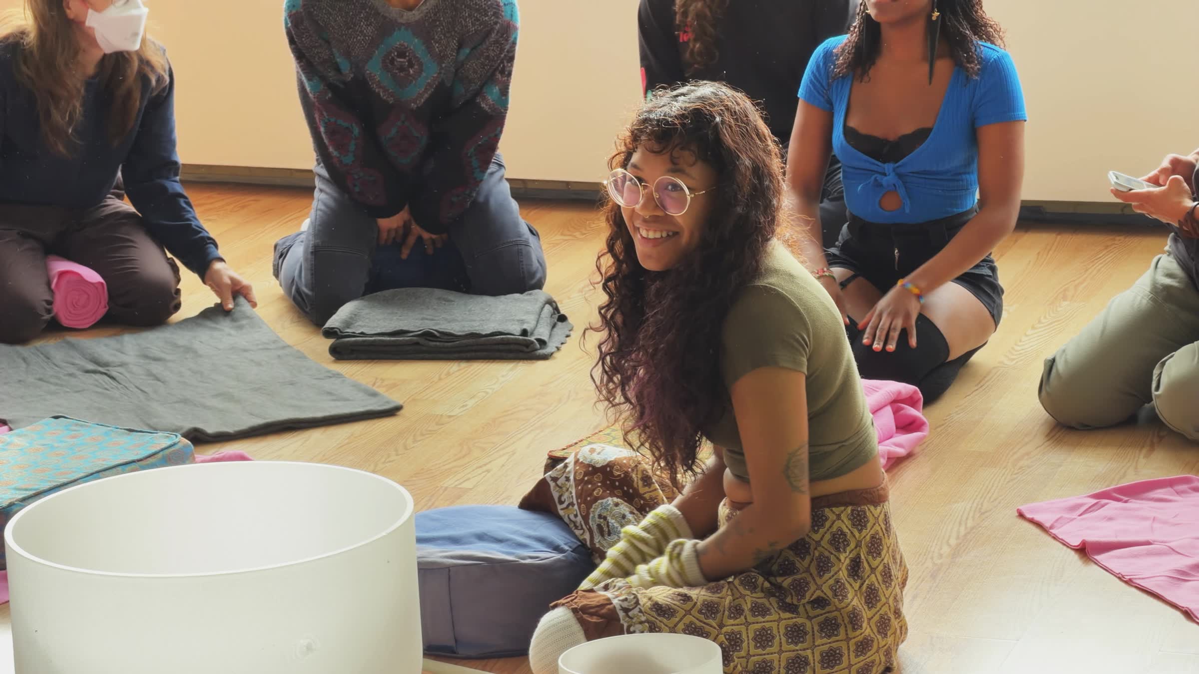 Group of people sitting on the floor with cushions and blankets, one smiling woman with glasses and gloves in the foreground.