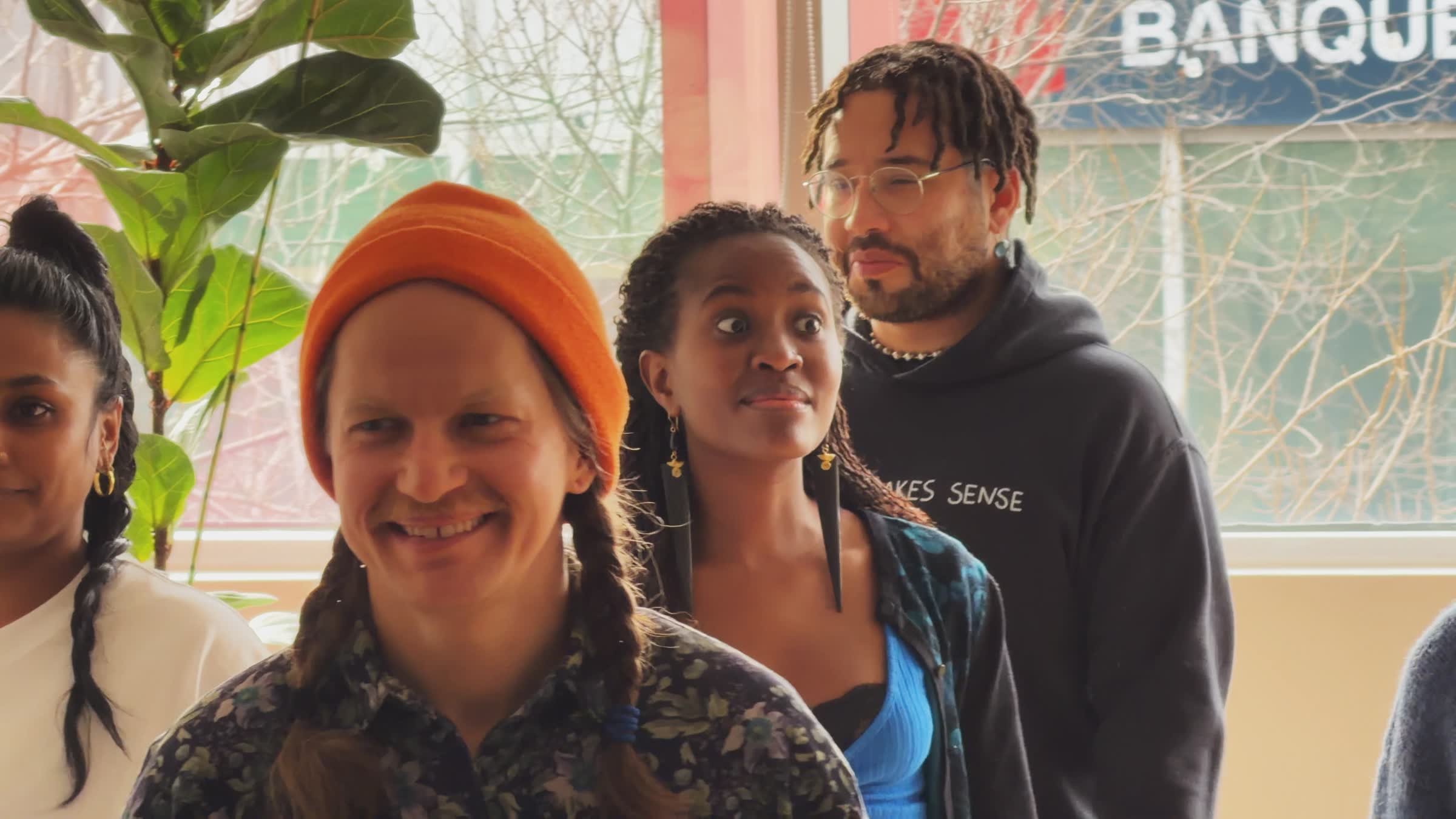 Four diverse young adults standing indoors near a window, one smiling wearing an orange beanie and two with braided hair.