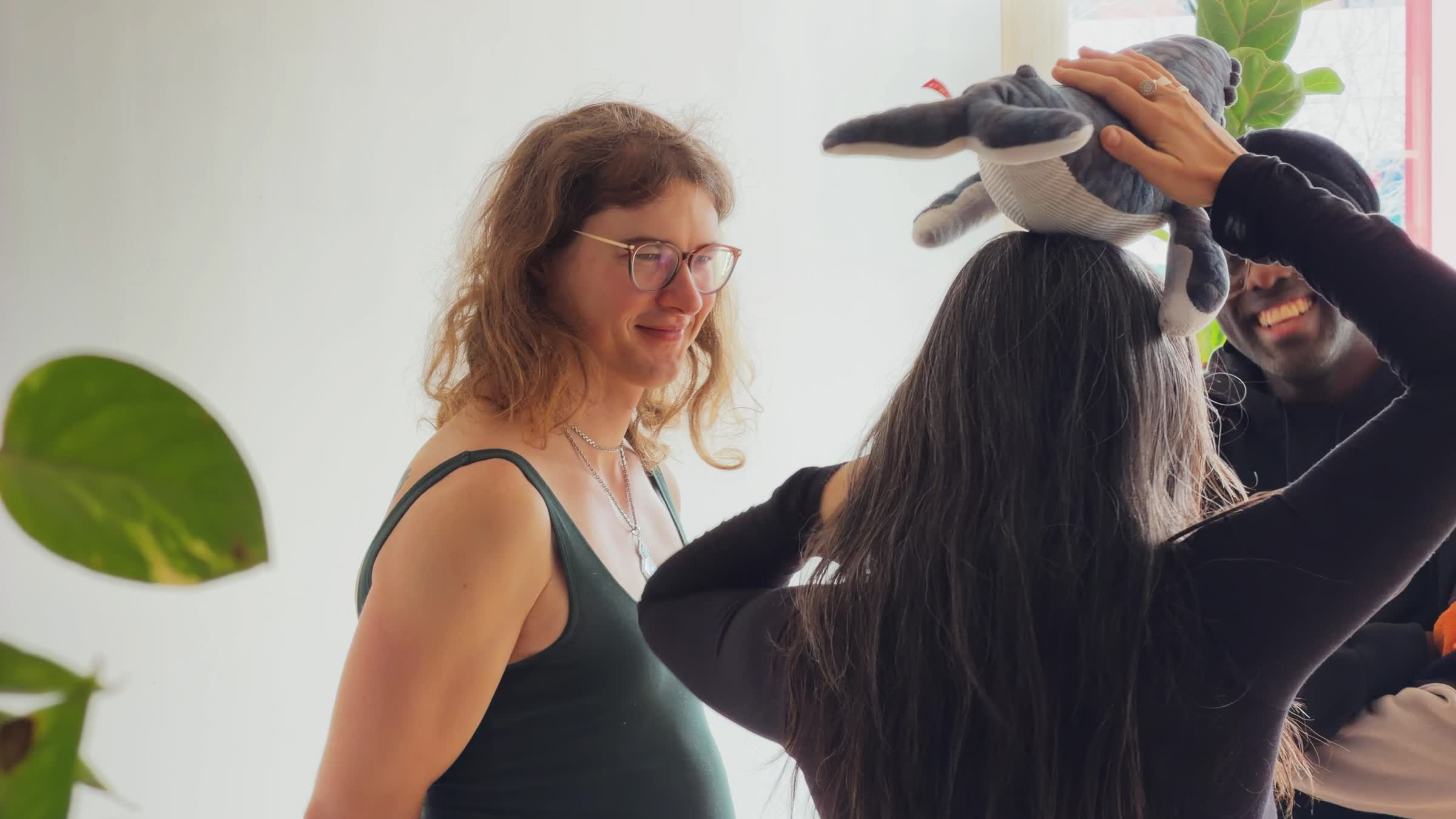 Three people indoors smiling and interacting, one person balancing a stuffed whale on their head.