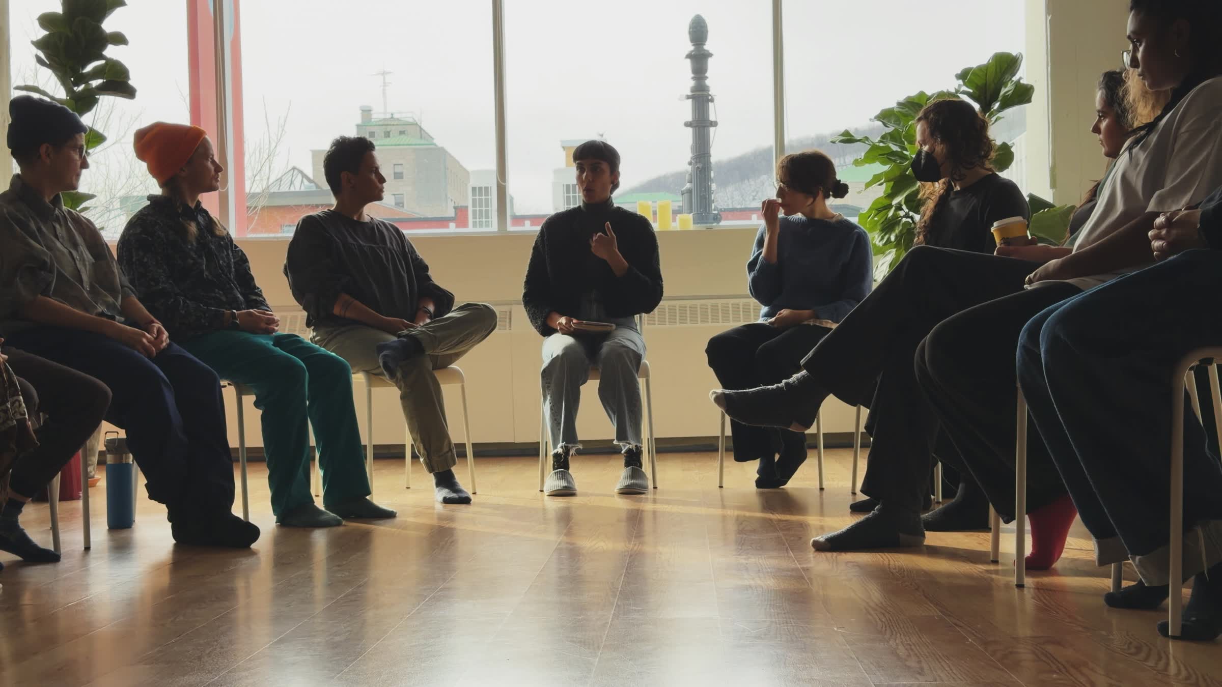 Group of people seated in a circle having a discussion in a room with large windows and wooden floor.