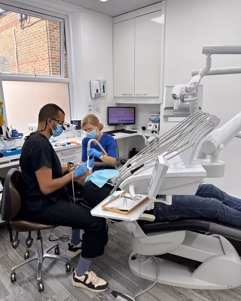A male dentist and female dental assistant wearing masks and gloves treating a patient lying in a dental chair in a modern clinic.