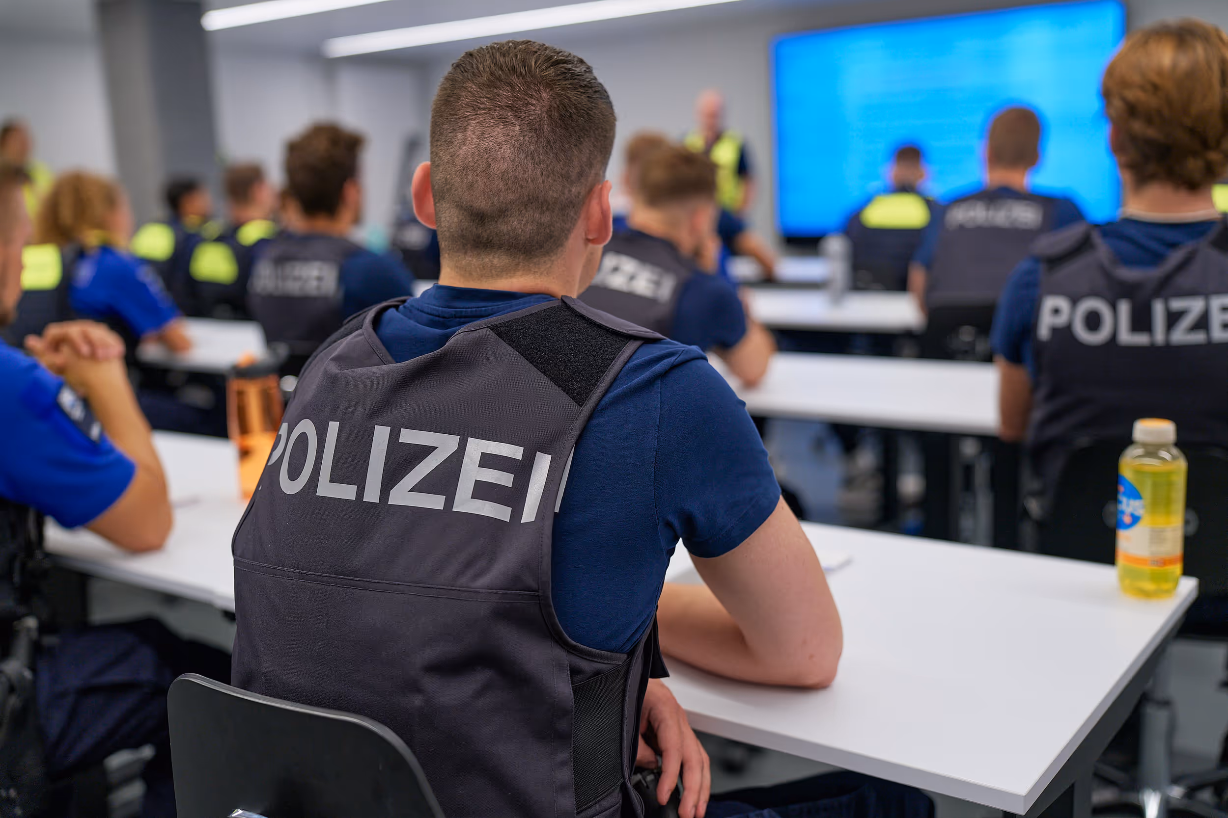 Polizeibeamte sitzen in einem Klassenzimmer und hören einer Person vor einem großen Bildschirm zu.