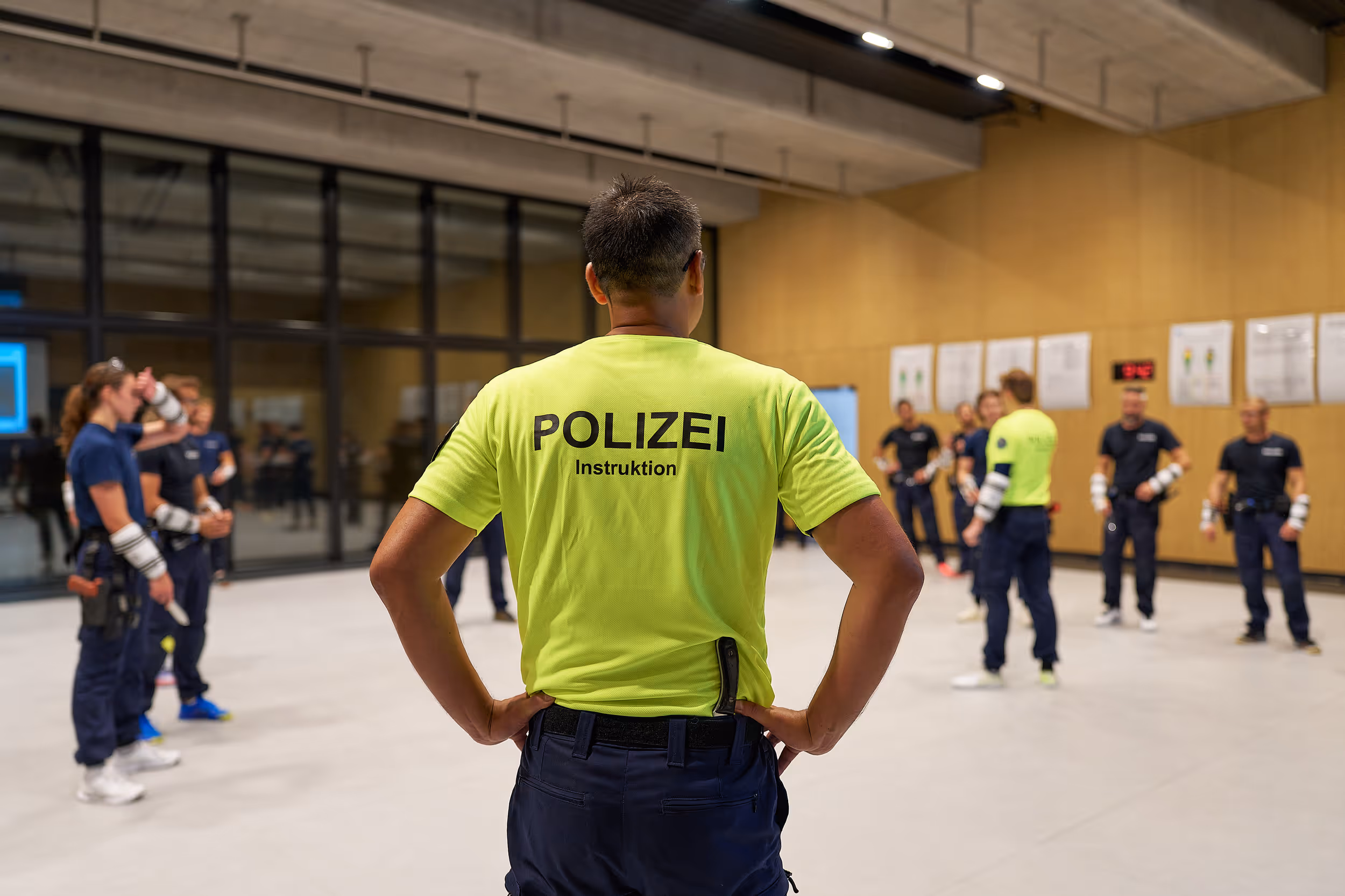 Polizeiausbilder in neongelbem T-Shirt hält Schulung für eine Gruppe von Polizisten in Trainingshalle.
