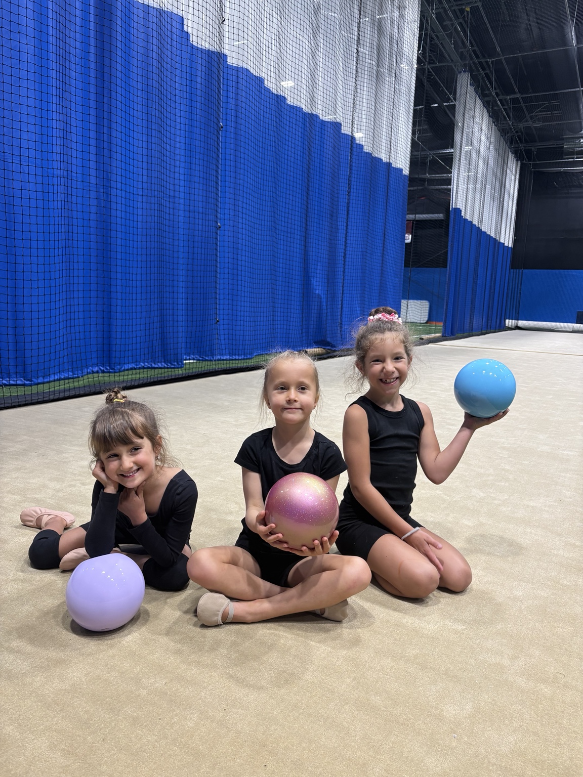 Three young Nord gymnasts sitting with rhythmic gymnastics balls during practice.