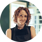 Friseurin mit lockigem Haar und schwarzer Schürze steht selbstbewusst vor dem Fenster.