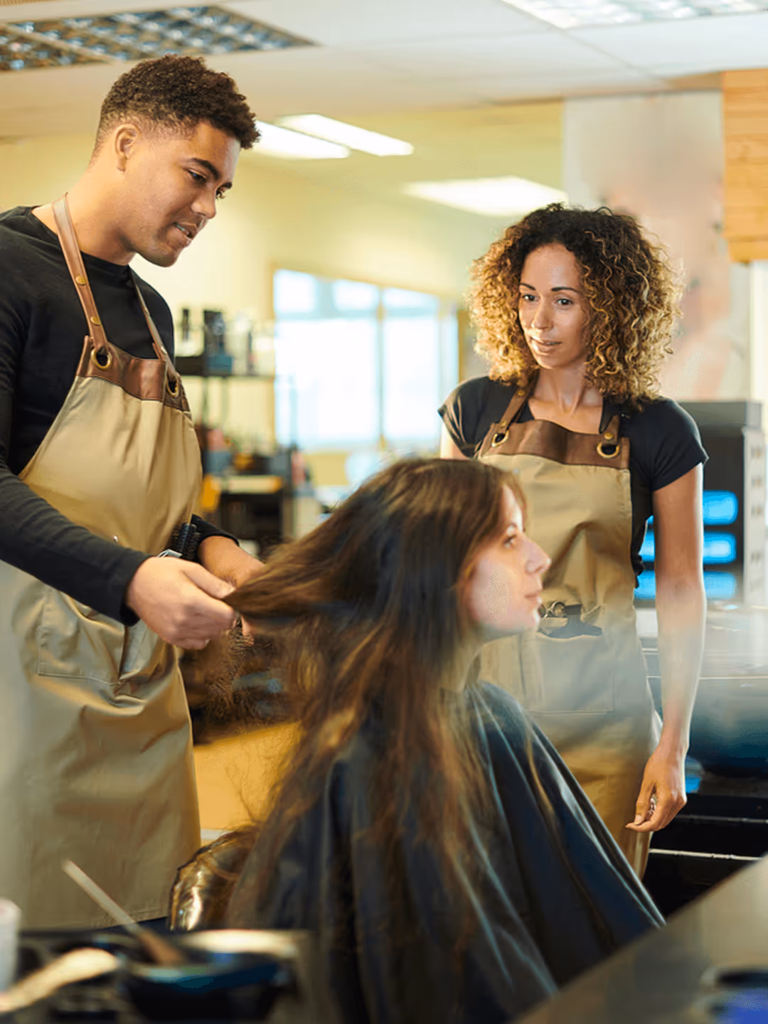 Männlicher Friseur föhnt die Haare einer Kundin, Kollegin steht daneben.
