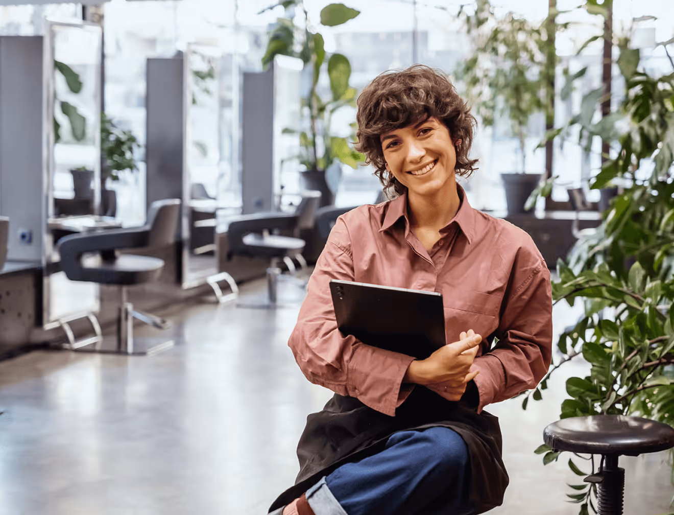 Ältere Friseurin sitzt lächelnd mit Tablet in einem hellen Salon.