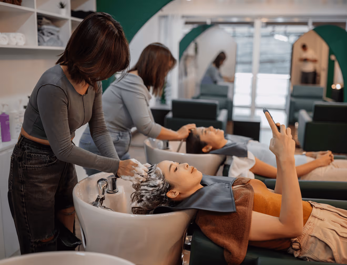 Kundin liegt mit nassem Haar am Waschbecken, Friseurin wäscht ihr die Haare.