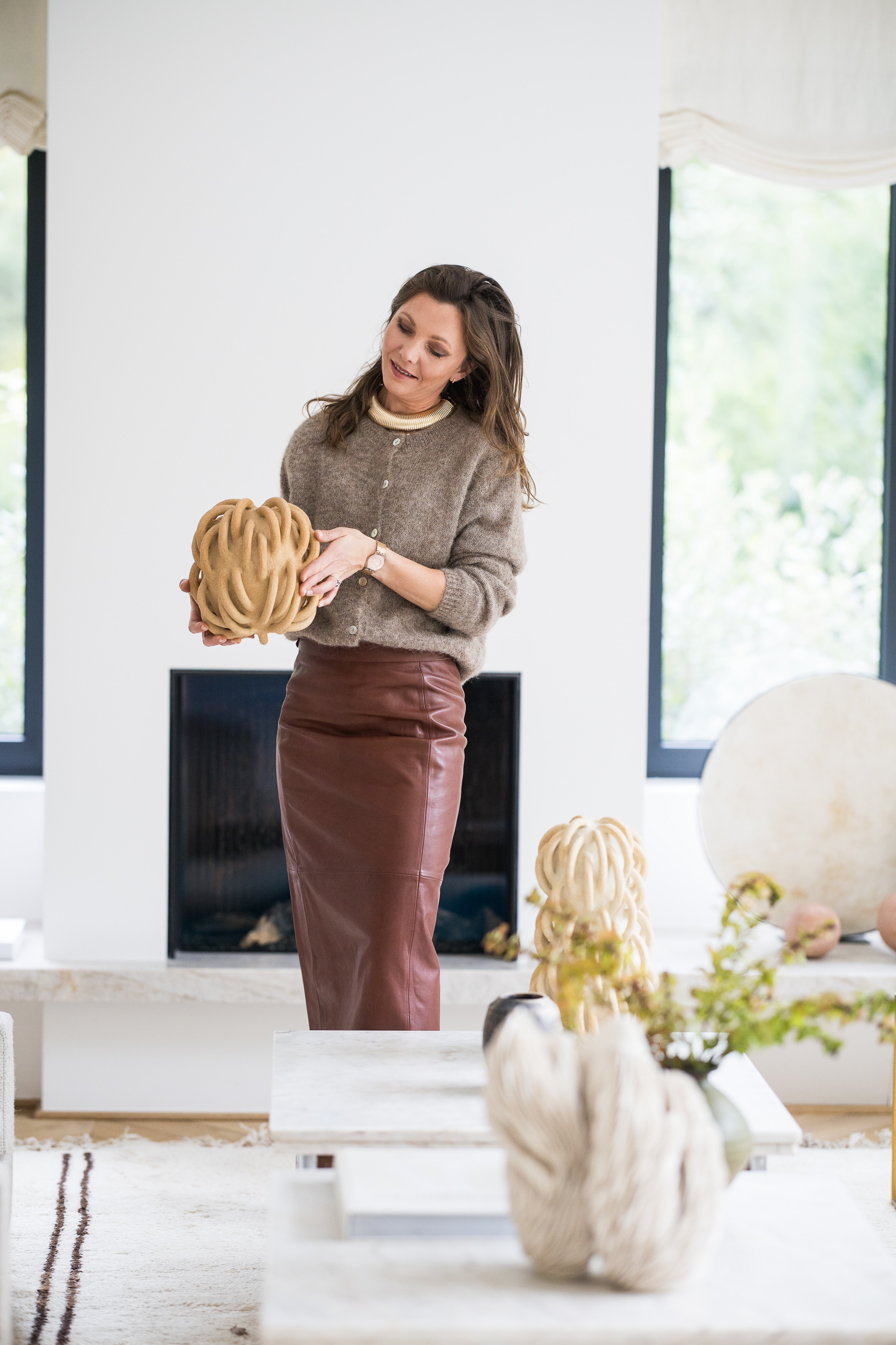 Alexia Werrie in a brown leather skirt and fuzzy sweater holding a tan sculptural object in a bright, modern living room.