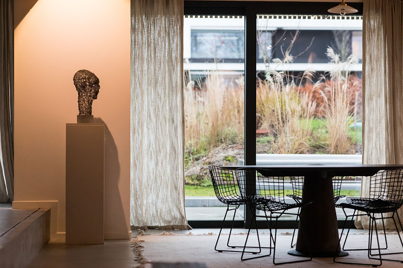 Modern interior with a wireframe human head sculpture on a pedestal next to a dining table and chairs by a large window with beige curtains.
