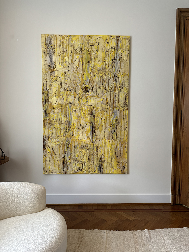 Abstract yellow and beige textured painting hanging on a white wall above a wooden floor and near a white curved chair.