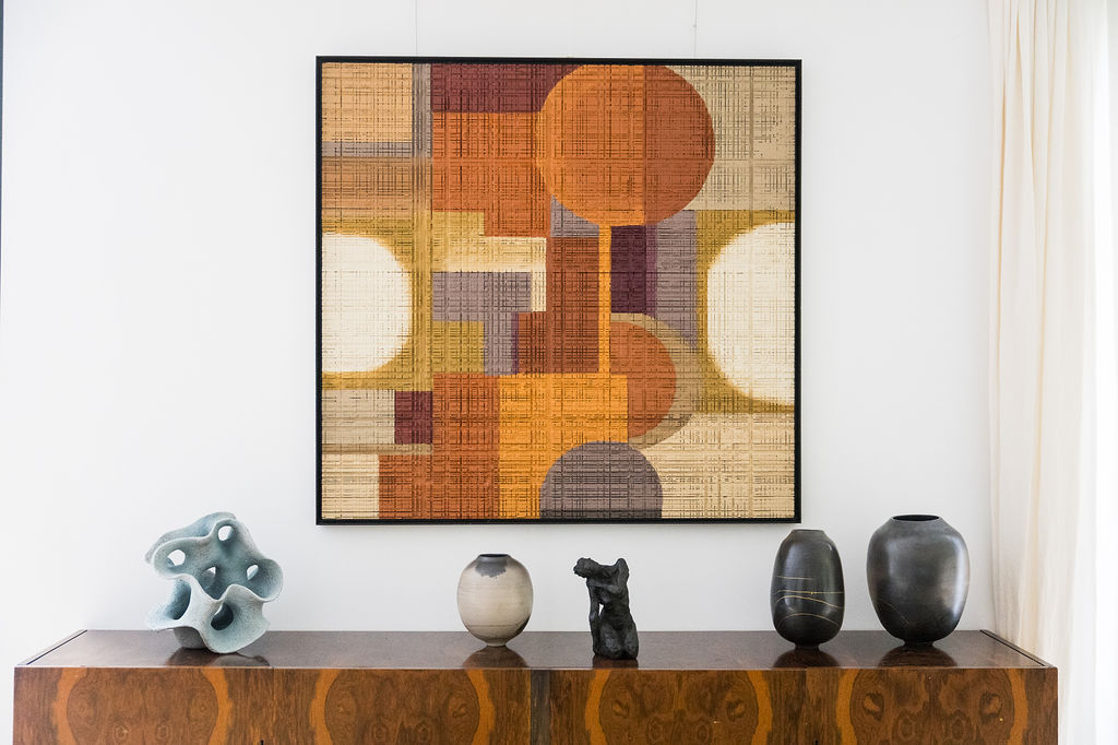 Abstract geometric artwork with warm tones hanging above a wooden cabinet displaying five sculptural vases and objects.