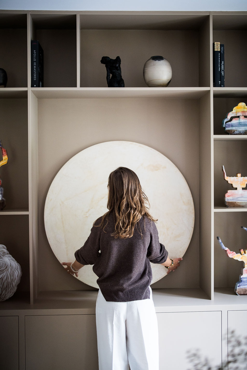 Woman with brown hair wearing a dark sweater and white pants, standing in front of a large round stone object on a beige shelving unit.