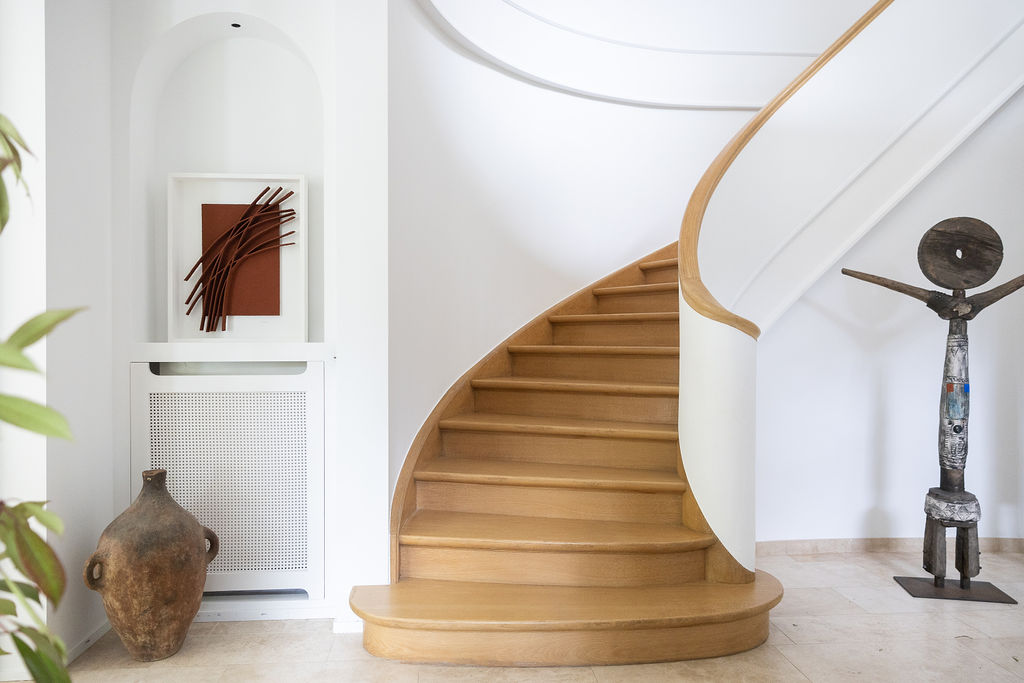 Curved wooden staircase with white railing in a minimalist interior featuring a large rustic vase, abstract wall art, and a tall wooden sculpture.