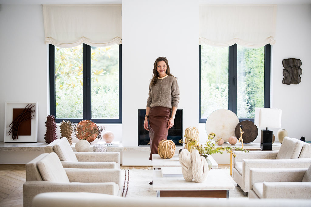 Woman standing and smiling in a bright, modern living room with beige armchairs, decorative vases, and large windows with cream curtains.