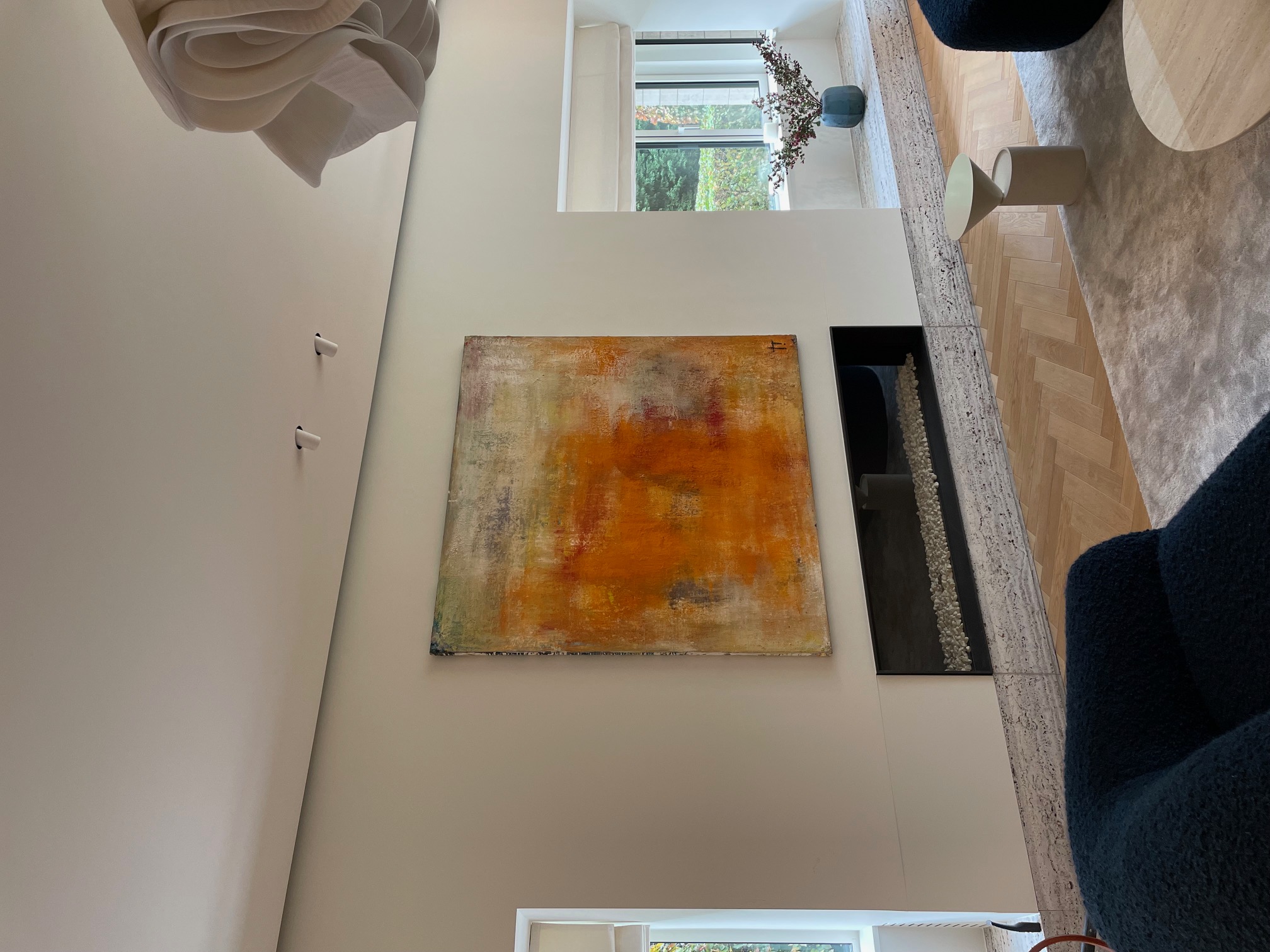 Modern living room with an abstract orange and beige painting above a sleek black fireplace, large windows with white blinds, and dark blue armchairs.