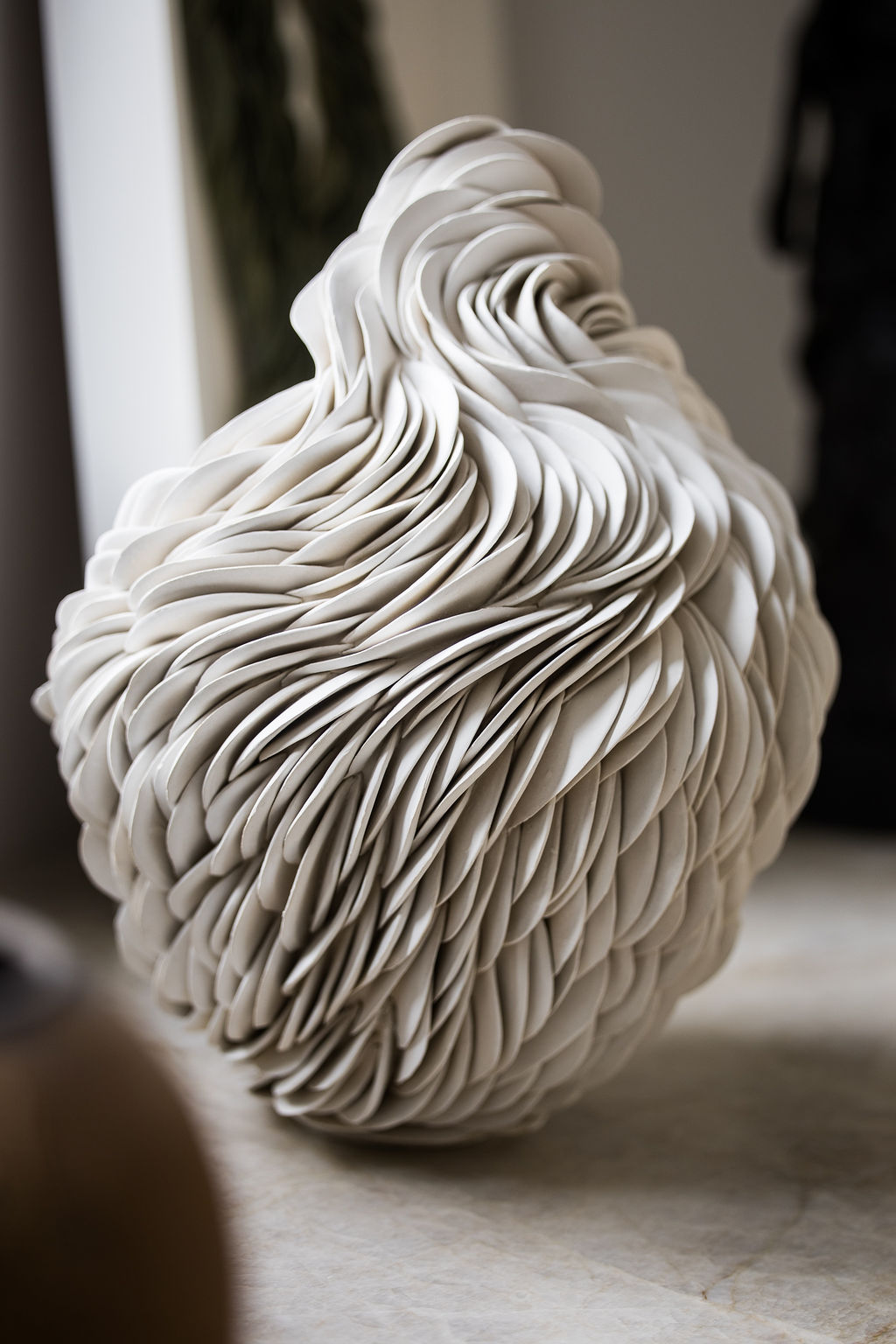 A sculptural white ceramic vase composed of hundreds of thin, hand-shaped clay layers that ripple and expand outward, creating a dynamic “distorted” silhouette. This one-of-a-kind piece embodies movement and light through its organic, undulating form
