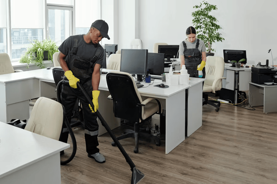 Zwei professionelle Reinigungskräfte in Arbeitskleidung und Handschuhen säubern ein helles und modernes Büro. Ein Mann saugt den Boden mit einem Nasssauger, während eine Frau im Hintergrund Oberflächen putzt.