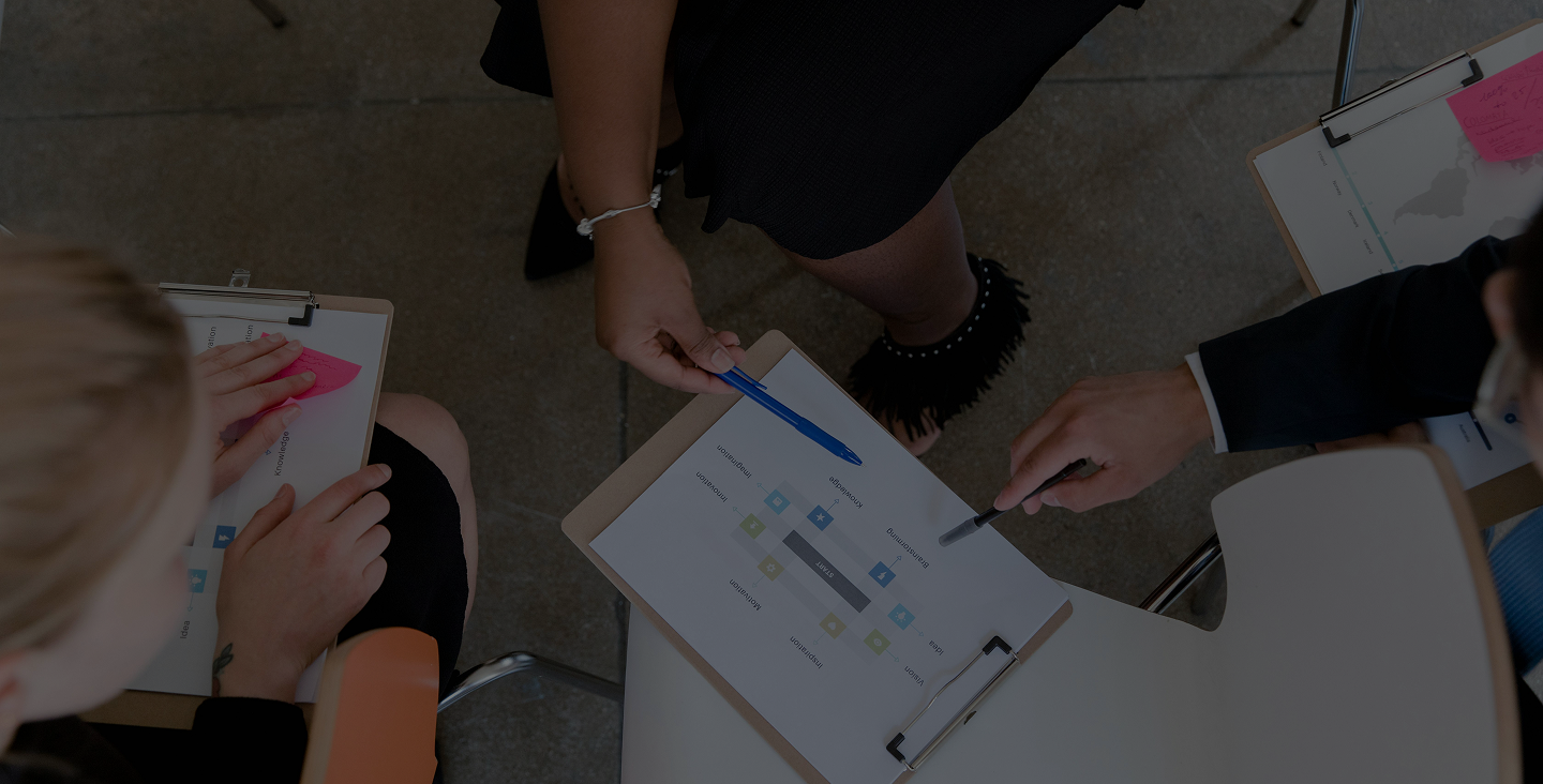 Three people seated in a circle holding clipboards with charts and sticky notes, discussing data with pens.