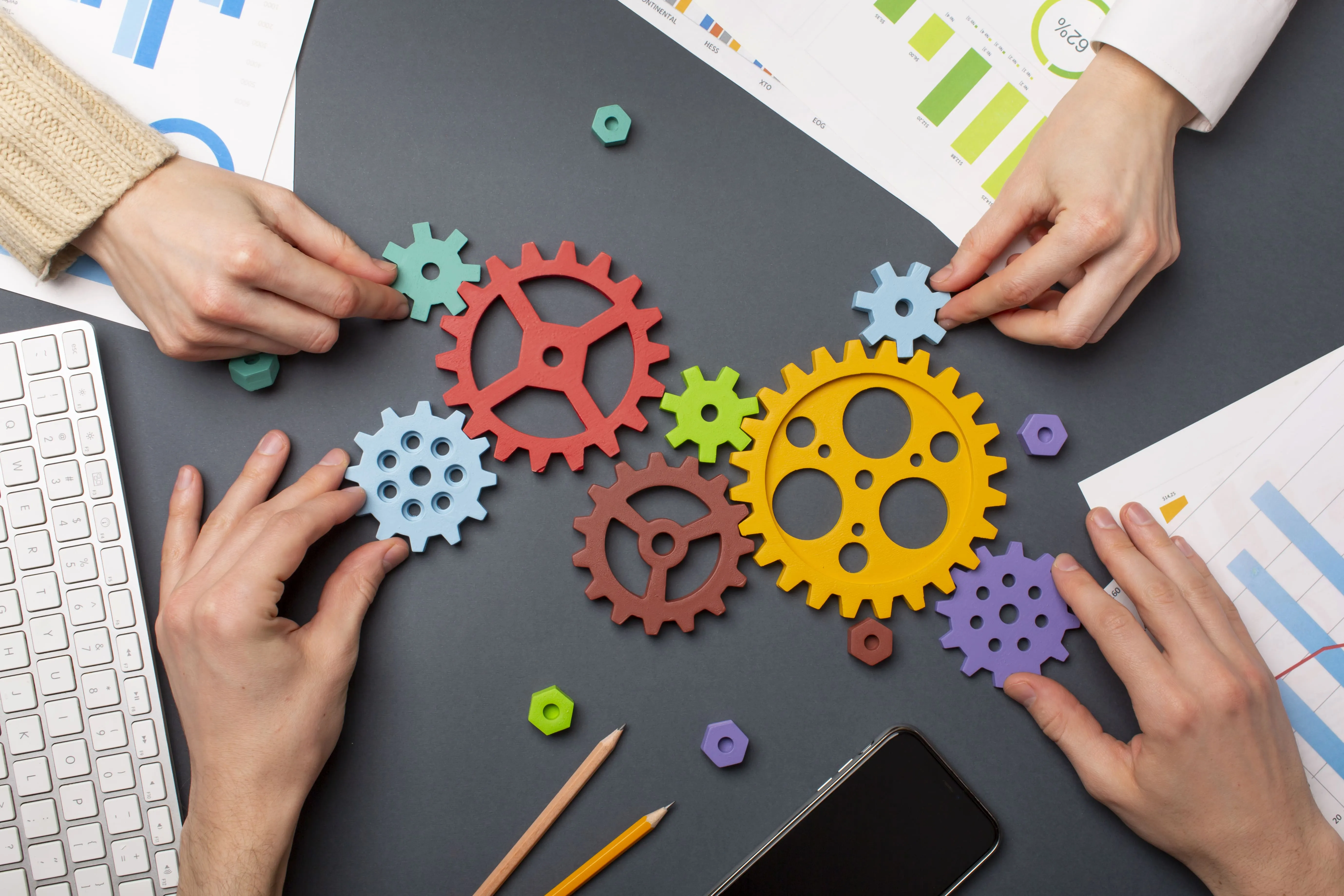 Four hands assembling colorful wooden gears on a gray desk with charts, pencils, keyboard, and smartphone.