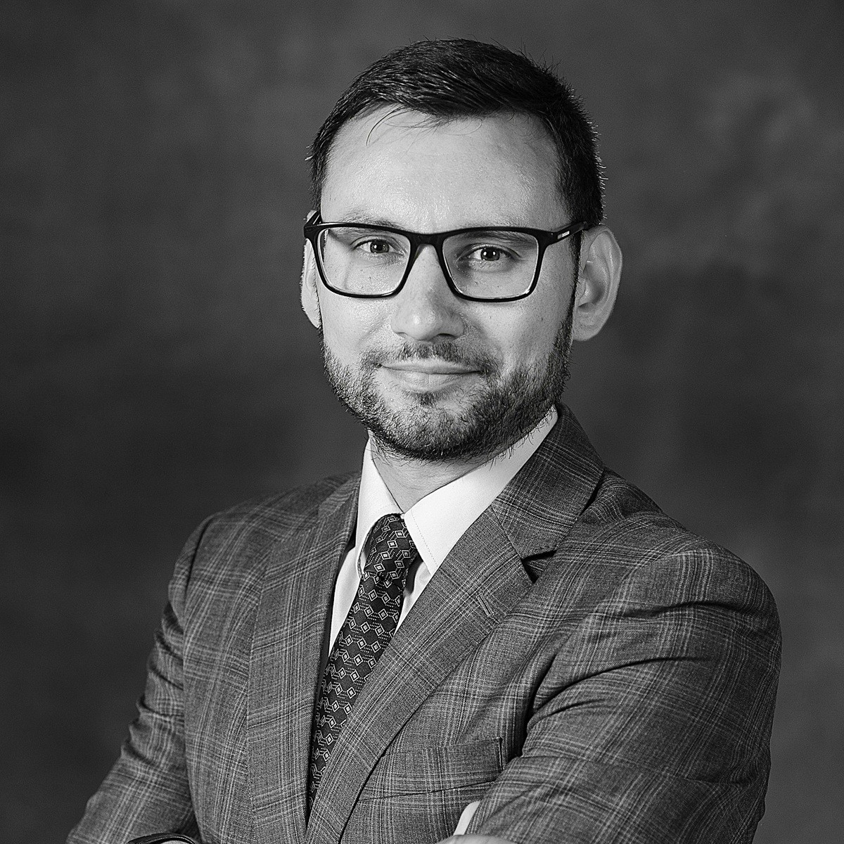 Man with glasses and beard in a plaid suit and patterned tie, standing with arms crossed against a dark background.